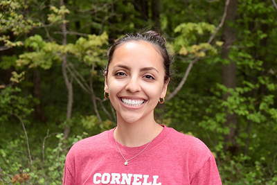 A smiling person wearing a Cornell shirt and a necklace with leafy trees behind her