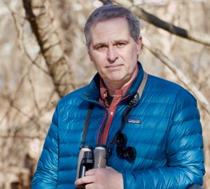 Outdoors, woods in the background, wearing blue coat and hold binoculars