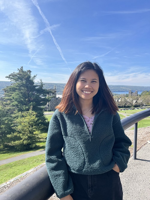 A smiling person with dark hair wearing a grey fleece and standing outdoors on Cornell Campus