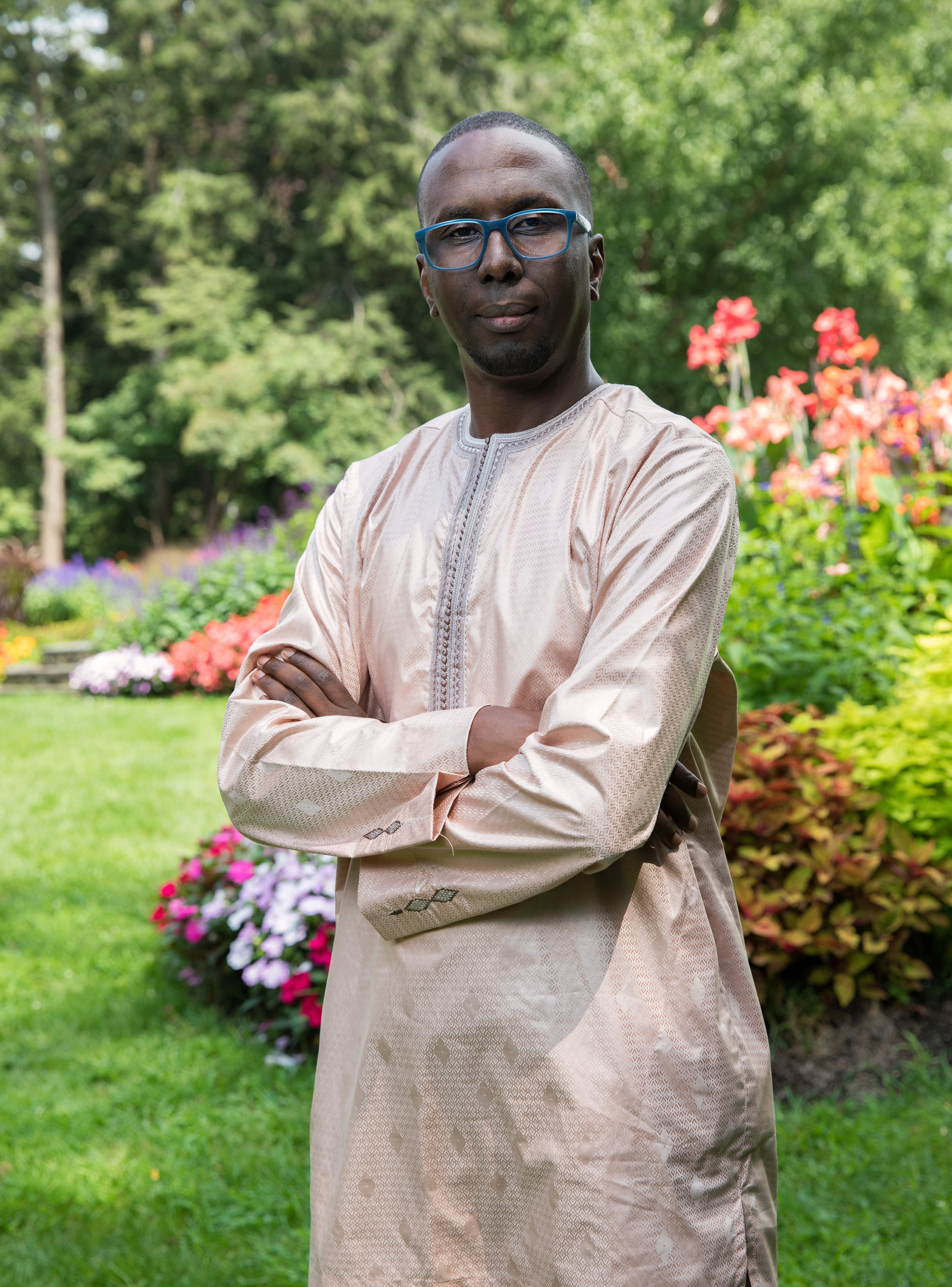 Oumar stands in a garden with his arms crossed. He is looking directly at the camera.