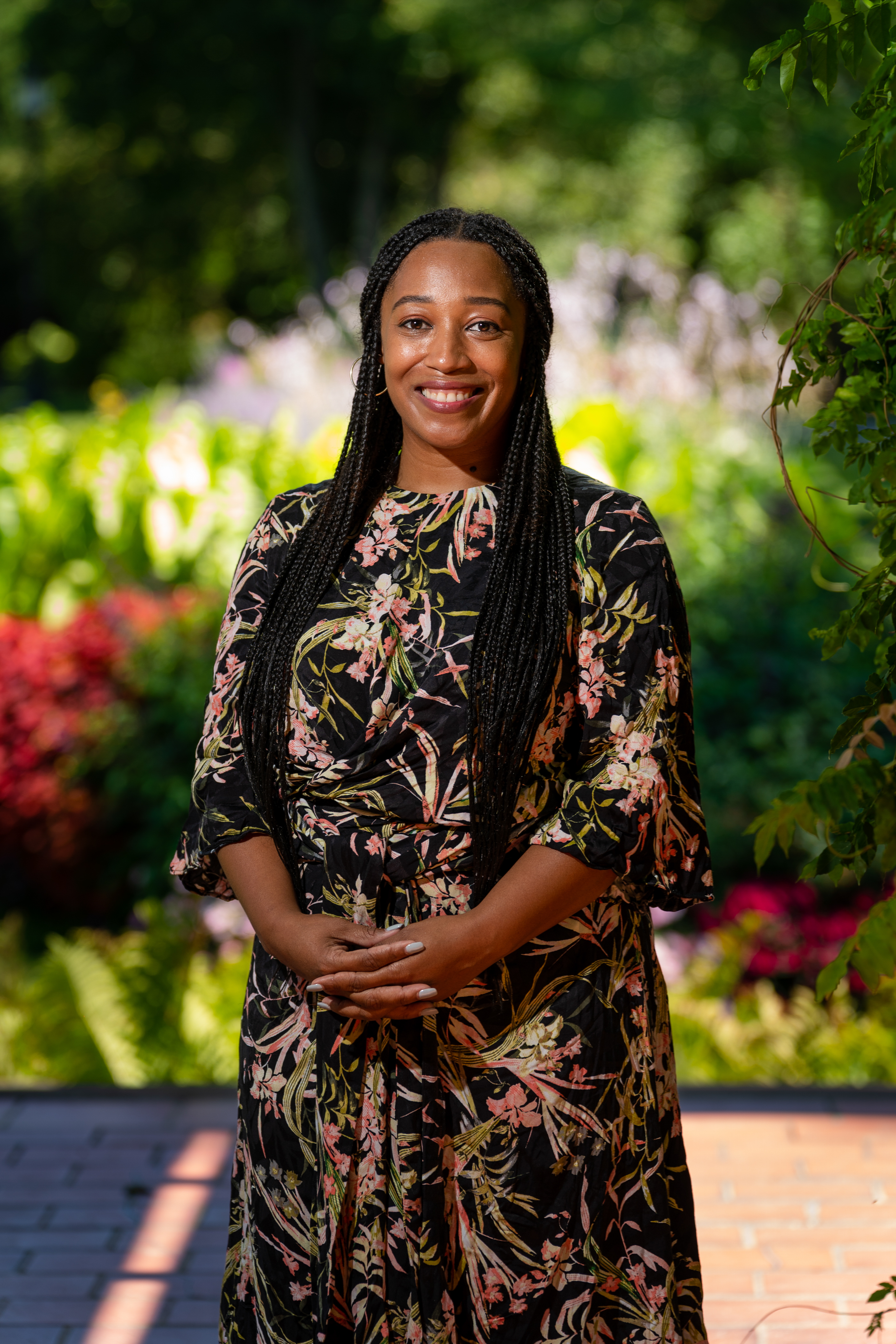 A smiling person with long braided hair stands outdoors in a garden wearing a floral dress.