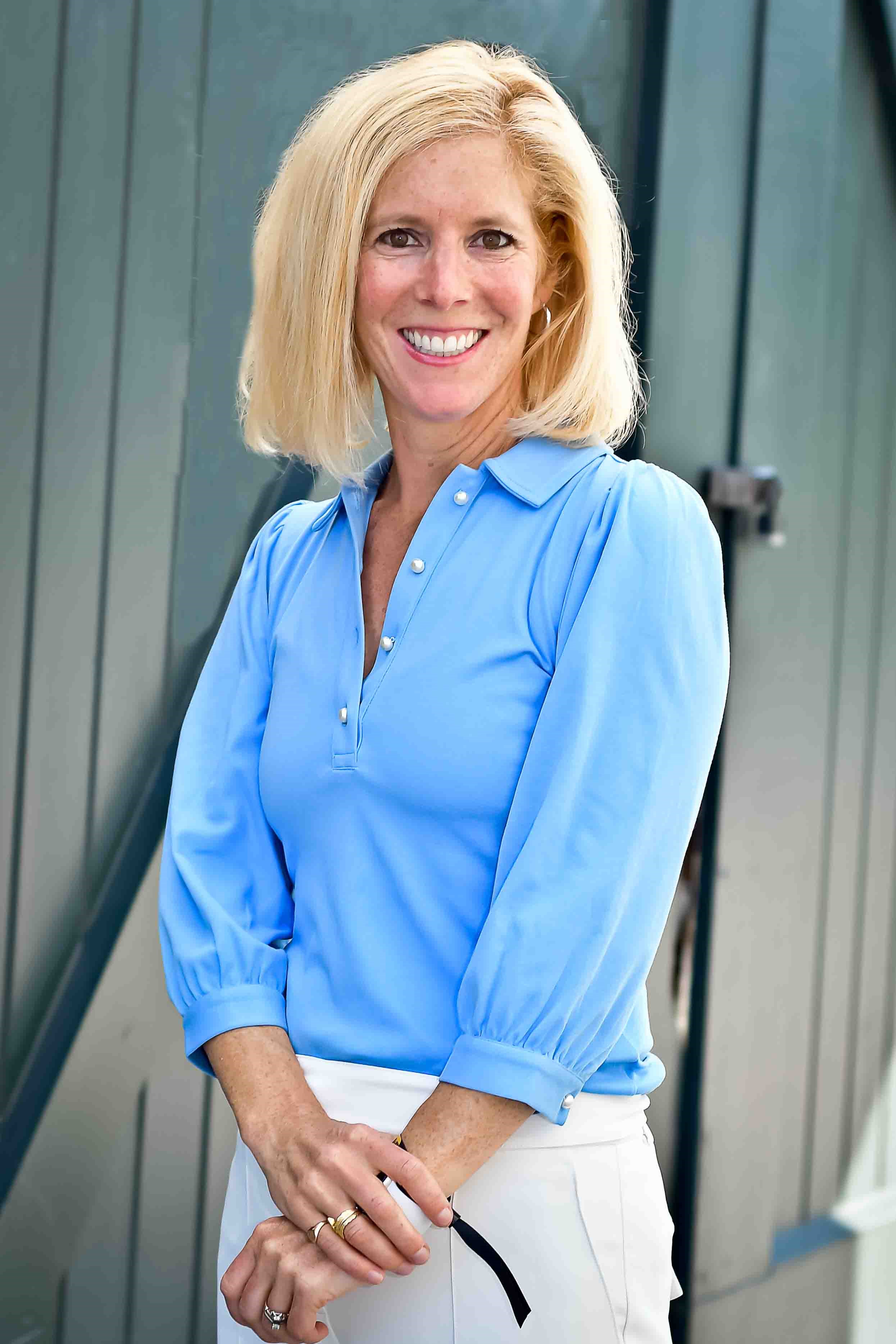 A smiling person with blond hair wearing a blue shirt standing outside.