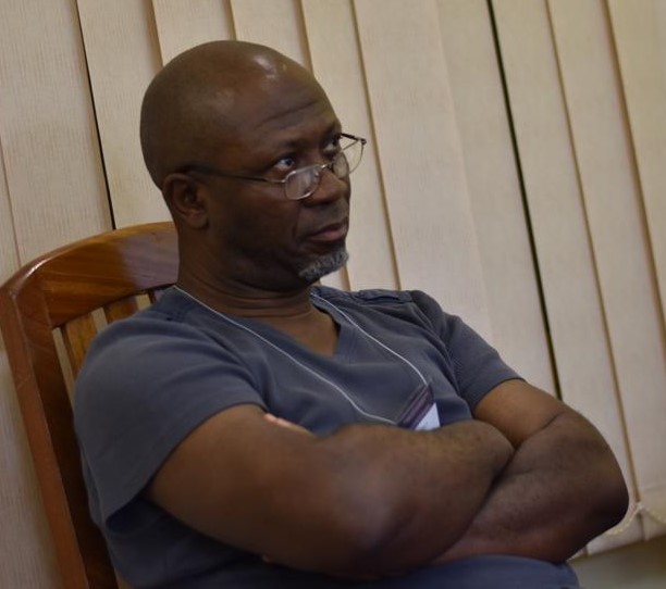 a three quarters angled picture of a bald man with glass, seated with his arms crossed in front of his chest.