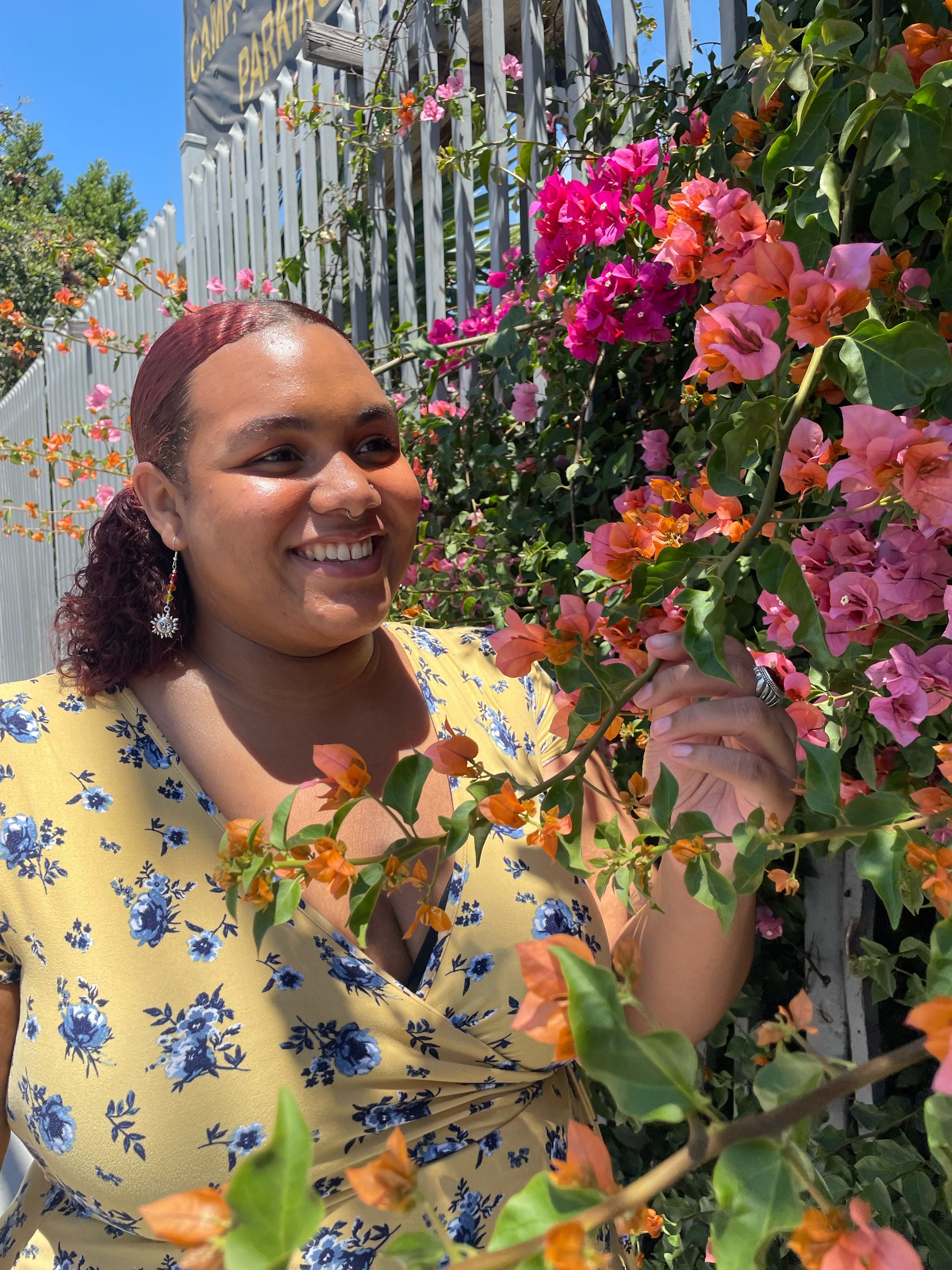 A person stands in a garden next to blooming flowers. She's wearing a floral dress and smiling.