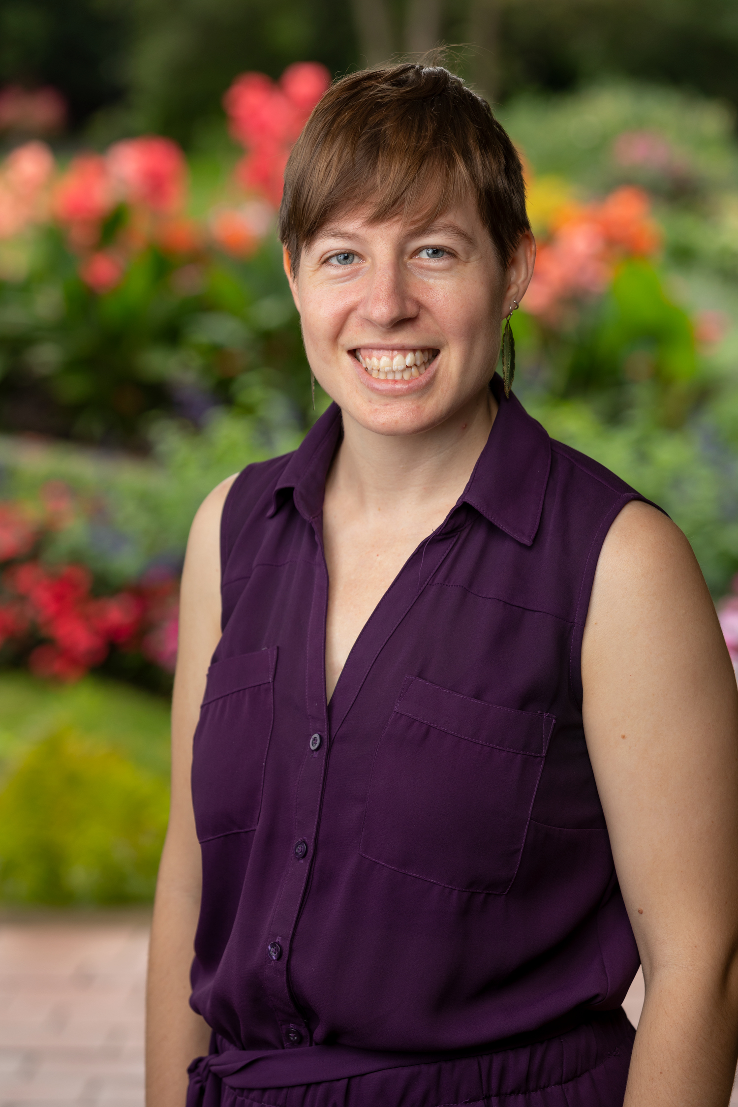 a smiling person in a purple shirt stands outside in front of a garden.