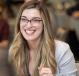 Jazlin smiling, long blond hair and glasses, informal setting, blurred background