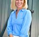 A smiling person with blond hair wearing a blue shirt standing outside.